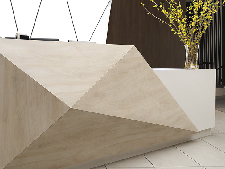 A modern geometric reception desk with a light beige wood grain finish and a white side pedestal holding a vase of yellow flowers, set in a contemporary interior.